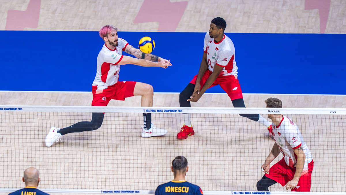 Polski siatkarz w białej koszulce z czerwonymi elementami i spodenkami, odbija piłkę do rozgrywającego. Jego kolega z drużyny, w podobnym stroju, przygotowuje się do zagrania. Na drugim planie widoczni są dwaj zawodnicy drużyny przeciwnej.