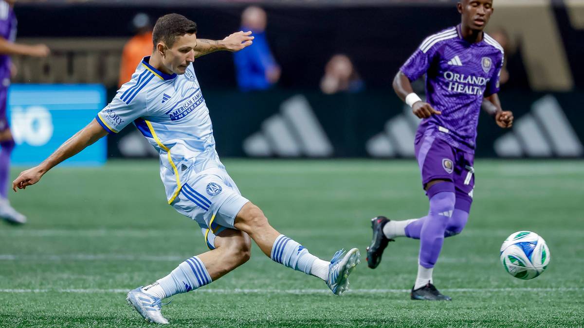 Dwóch piłkarzy na zielonym boisku. Jeden w jasnoniebiesko-białym stroju kopie piłkę, drugi w fioletowym stroju biegnie w kierunku piłki.