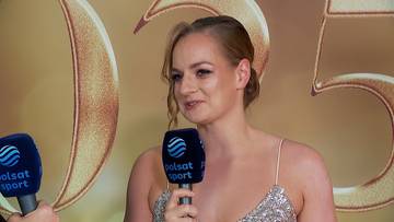 Kobieta z krótkimi, blond włosami, ubrana w błyszczącą sukienkę, trzymająca mikrofon, przemawiająca do dziennikarzy.