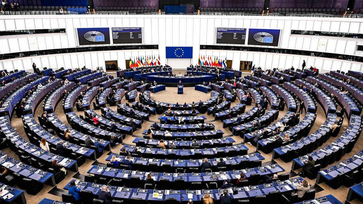 Widok z góry na salę plenarną Parlamentu Europejskiego z rzędami niebieskich siedzeń i centralną mównicą z flagą UE.