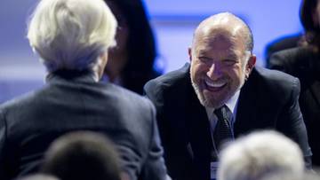 Mężczyzna w garniturze śmieje się podczas spotkania, obok siedzi osoba z siwymi włosami odwr&oacute;cona plecami.