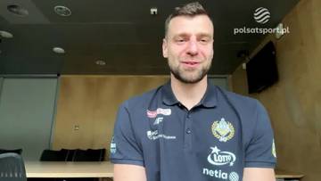 Kamil Łączyński, zawodnik koszykówki, ubrany w koszulkę klubową z widocznymi logotypami, uśmiecha się do kamery.