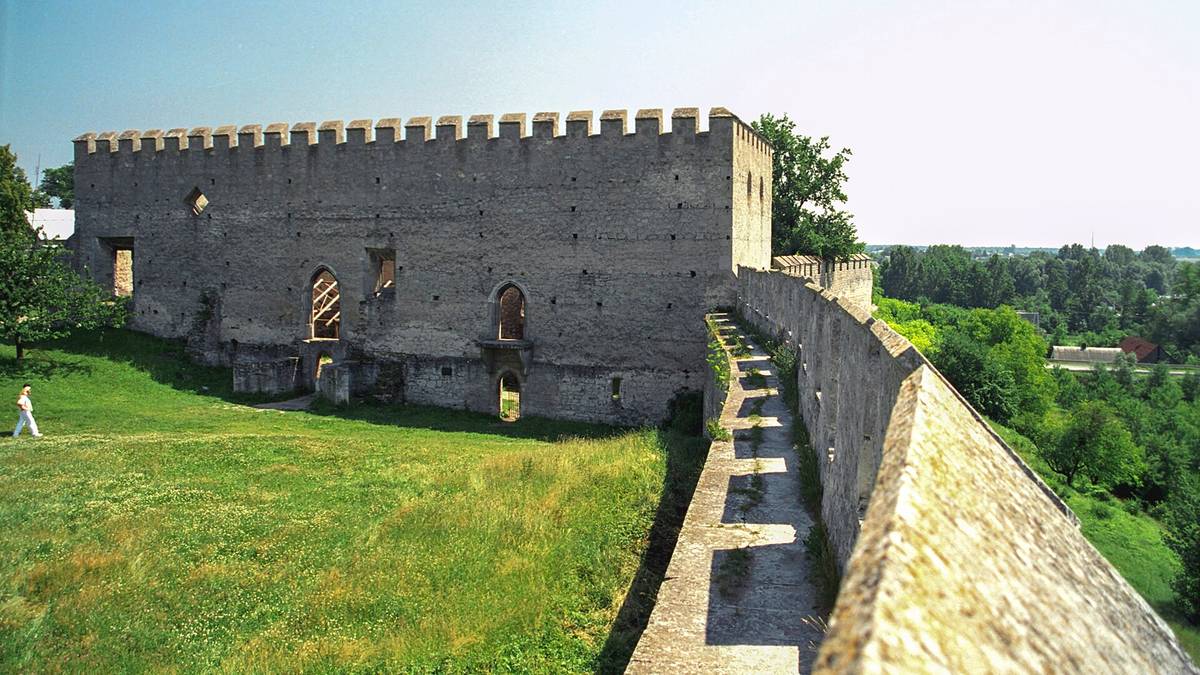 Fragment średniowiecznych murów obronnych i kamiennego budynku na wzgórzu, z widokiem na zieloną dolinę.