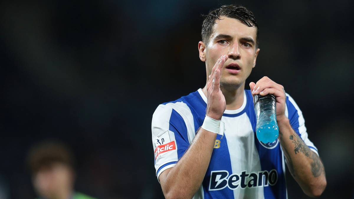 Piłkarz w białej koszulce z niebieskimi pasami, trzymający w ręku niebieską butelkę z napojem izotonicznym. W tle rozmyty tłum.