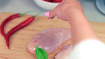 Ręka doprawia surowe piersi kurczaka na desce z chili i ziołami.