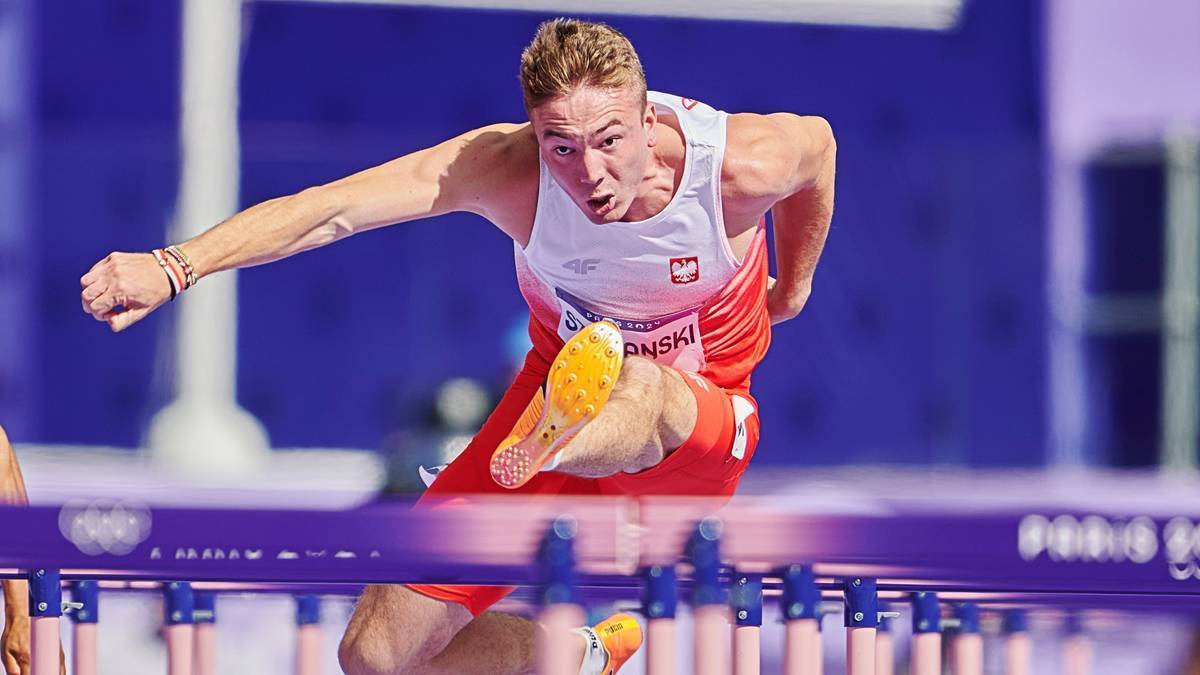 Jakub Szymański w białej koszulce z napisem Polska i czerwonymi spodenkami pokonuje płotek podczas biegu na 100 metrów.