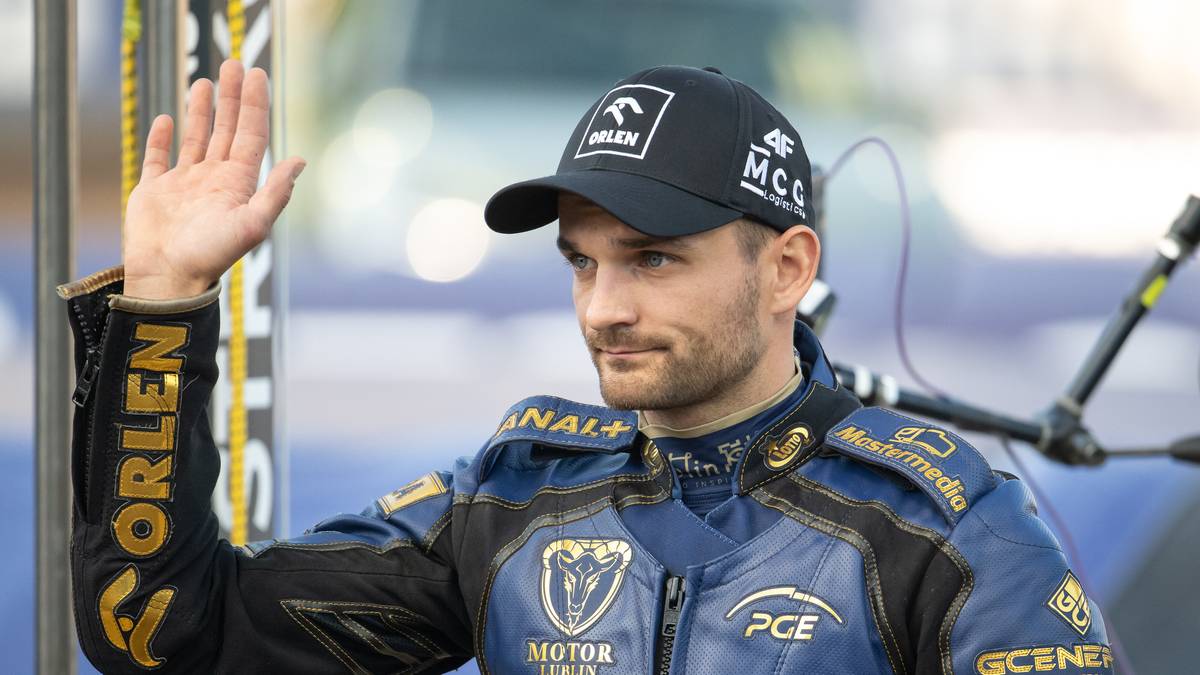 Bartosz Zmarzlik w stroju żużlowym i czapce z daszkiem ORLEN, macha prawą ręką na powitanie.