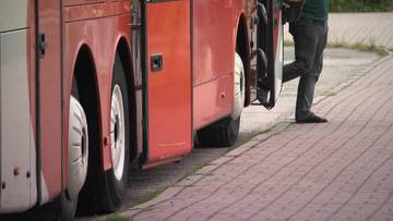 Fragment czerwonego autobusu z otwartymi drzwiami i pasażerem stojącym przy chodniku z kostki brukowej.