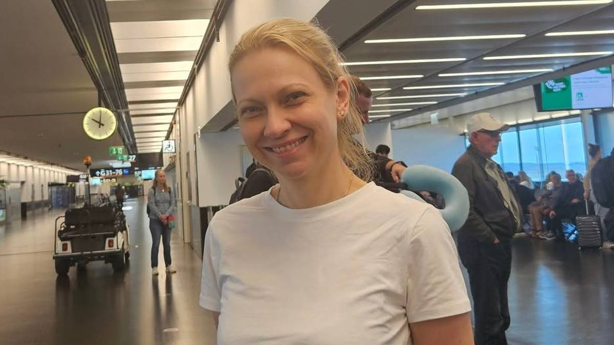 Uśmiechnięta kobieta z blond włosami w białej koszulce stoi w tle lotniska.