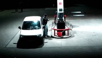 Mężczyzna stoi przy dystrybutorze paliwa na stacji nocą, obok stoi biały samoch&oacute;d.