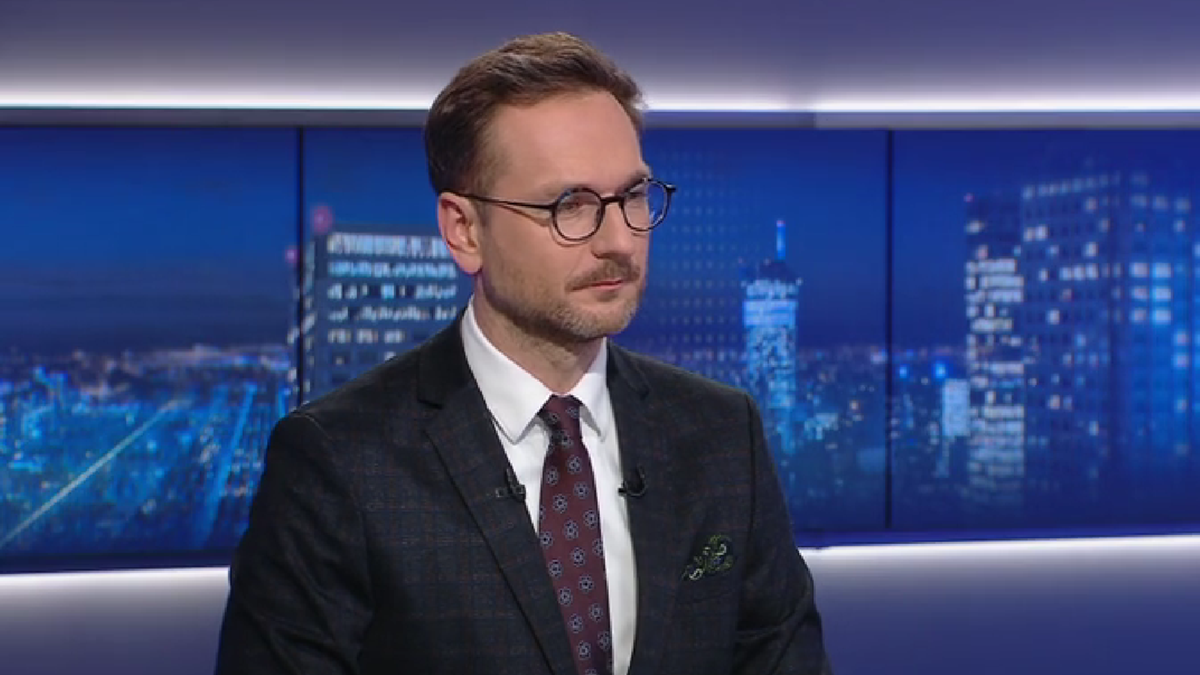 Mężczyzna w okularach i garniturze siedzi przed ekranami przedstawiającymi nocną panoramę miasta.