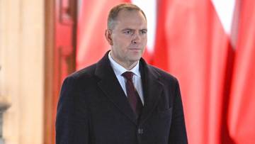 Mężczyzna w garniturze i płaszczu, stojący przed czerwonym tłem.