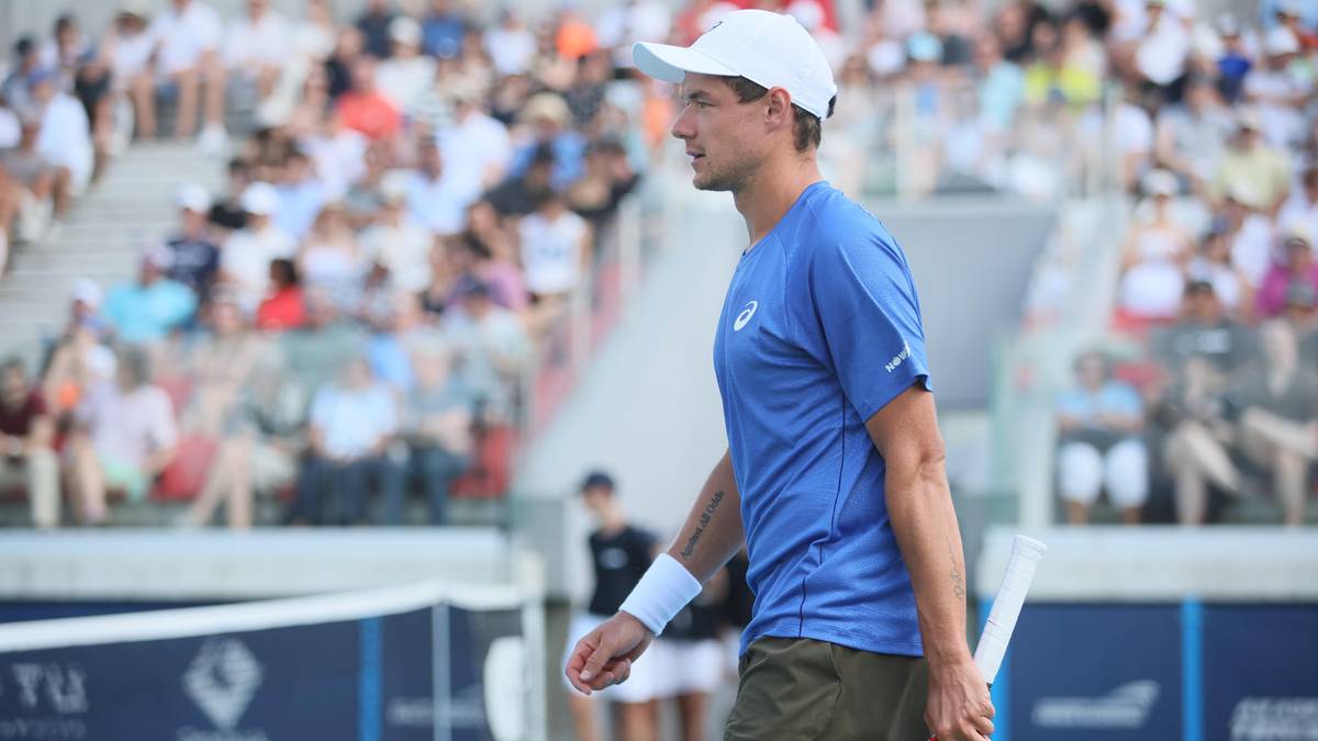 Tenisista w niebieskiej koszulce i białej czapce na korcie tenisowym, obr&oacute;cony w prawo, z rakietą w dłoni. Na drugim planie widownia.