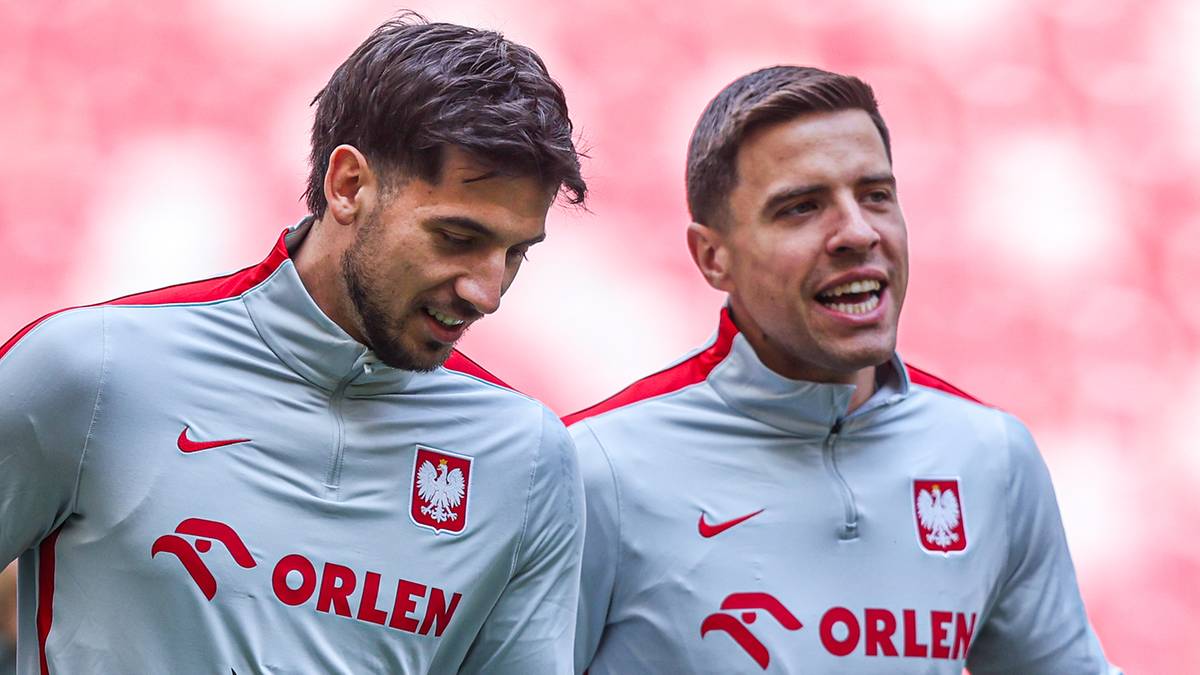 Piłkarze w szarych strojach treningowych z herbem Polski i logiem sponsora Orlen, biegną na boisku stadionu.