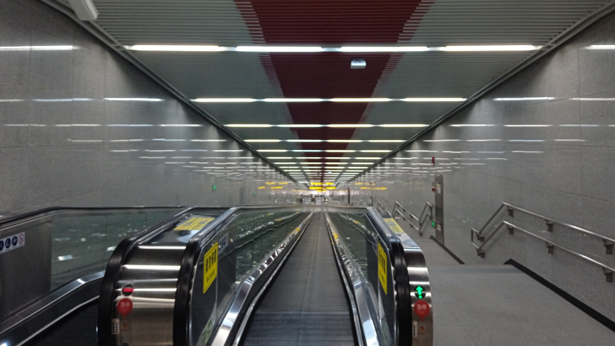 Długi korytarz stacji metra z ruchomymi schodami i schodami po prawej stronie. Sufit jest wyłożony panelami z oświetleniem i ozdobiony czerwonym wzorem.