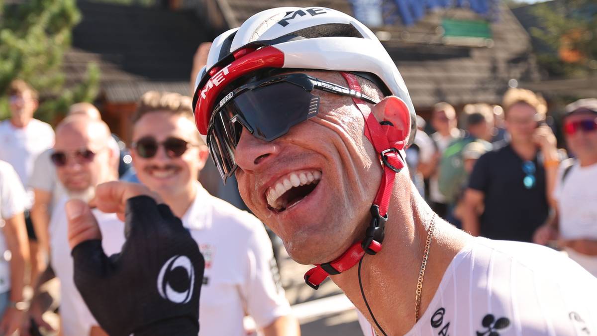 Zbliżenie na twarz Rafała Majki w kasku i okularach rowerowych, uśmiechniętego podczas wyścigu kolarskiego.