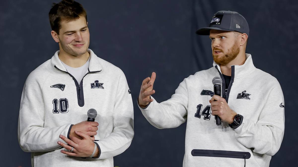 Dw&oacute;ch mężczyzn w białych bluzach z logo Super Bowl LVII, jeden z nich trzyma mikrofon i gestykuluje, drugi patrzy na niego z zaciekawieniem.