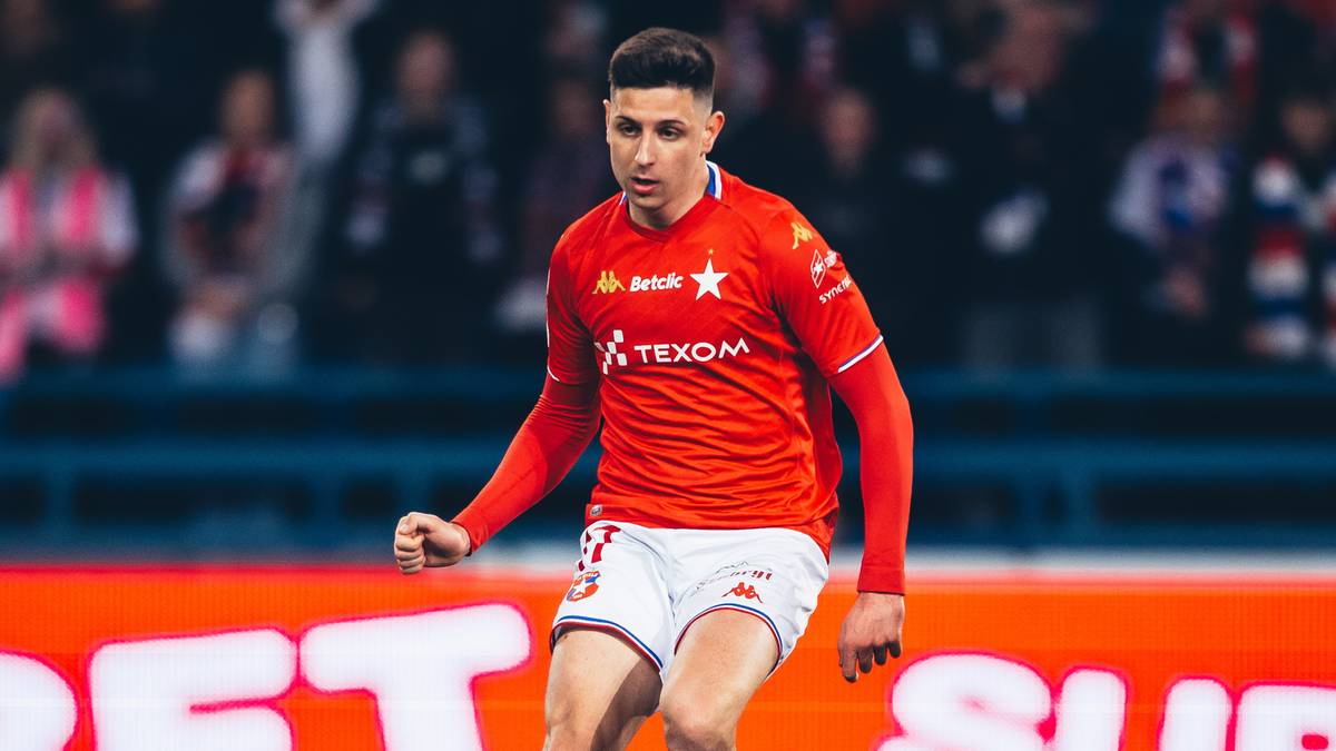 Piłkarz w czerwonej koszulce Wisły Kraków z numerem 10 na plecach biegnie z piłką na murawie stadionu.