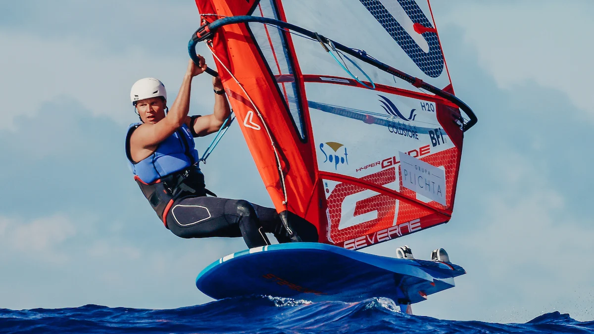 Mężczyzna na desce windsurfingowej z żaglem w powietrzu nad wodą.