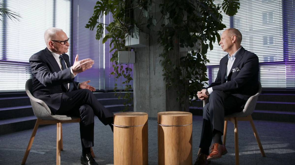 Dw&oacute;ch mężczyzn w eleganckich garniturach siedzi na krzesłach w nowoczesnym wnętrzu i rozmawia. Jeden z mężczyzn gestykuluje dłońmi podczas wypowiedzi.