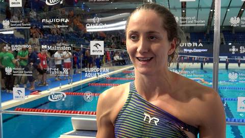 Katarzyna Wasick, uśmiechnięta pływaczka w stroju TYR, udziela wywiadu na tle basenu pływackiego.
