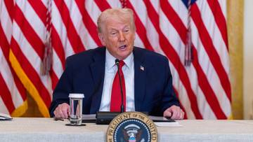 Donald Trump siedzi przy stole z amerykańskimi flagami w tle.