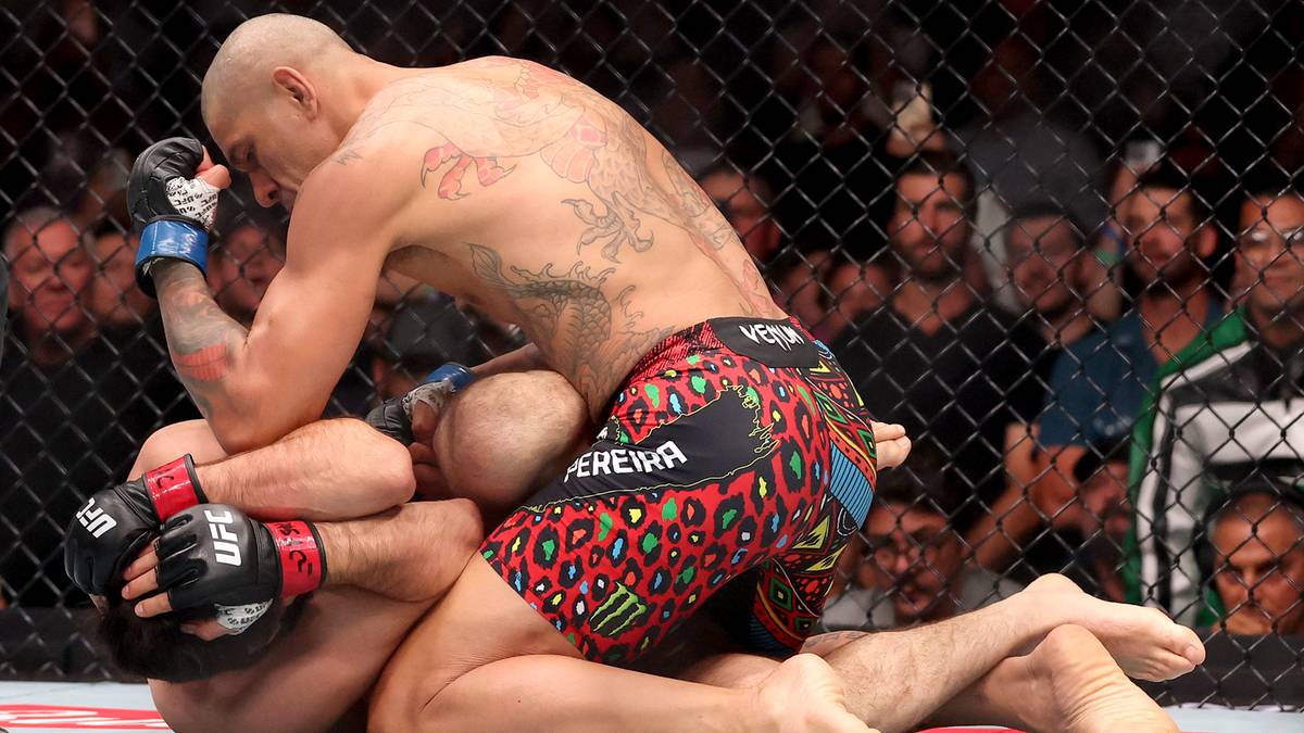 Zawodnik MMA w kolorowych spodenkach z czerwonymi i czarnymi wzorami klęczy nad przeciwnikiem, zadając uderzenie. Widownia w tle.