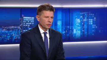 Mężczyzna w ciemnym garniturze i fioletowym krawacie siedzi, patrząc na wprost. W tle widoczny jest ekran z rozmazanym widokiem nocnego miasta.