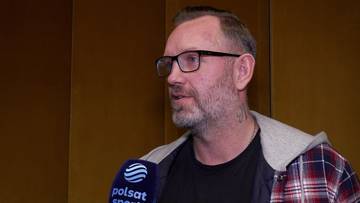 Grzegorz Tkaczyk w okularach i z zarostem, ubrany w kratę koszulę, z mikrofonem Polsat Sport przed twarzą.