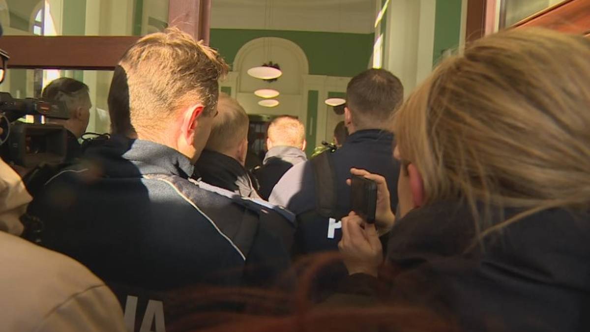 Policjanci wchodzący do budynku, w tle widoczni inni funkcjonariusze i cywilne osoby.