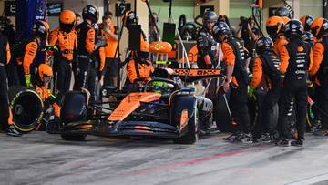 Zespół mechaników w pomarańczowych strojach otacza bolid Formuły 1 w boksie.