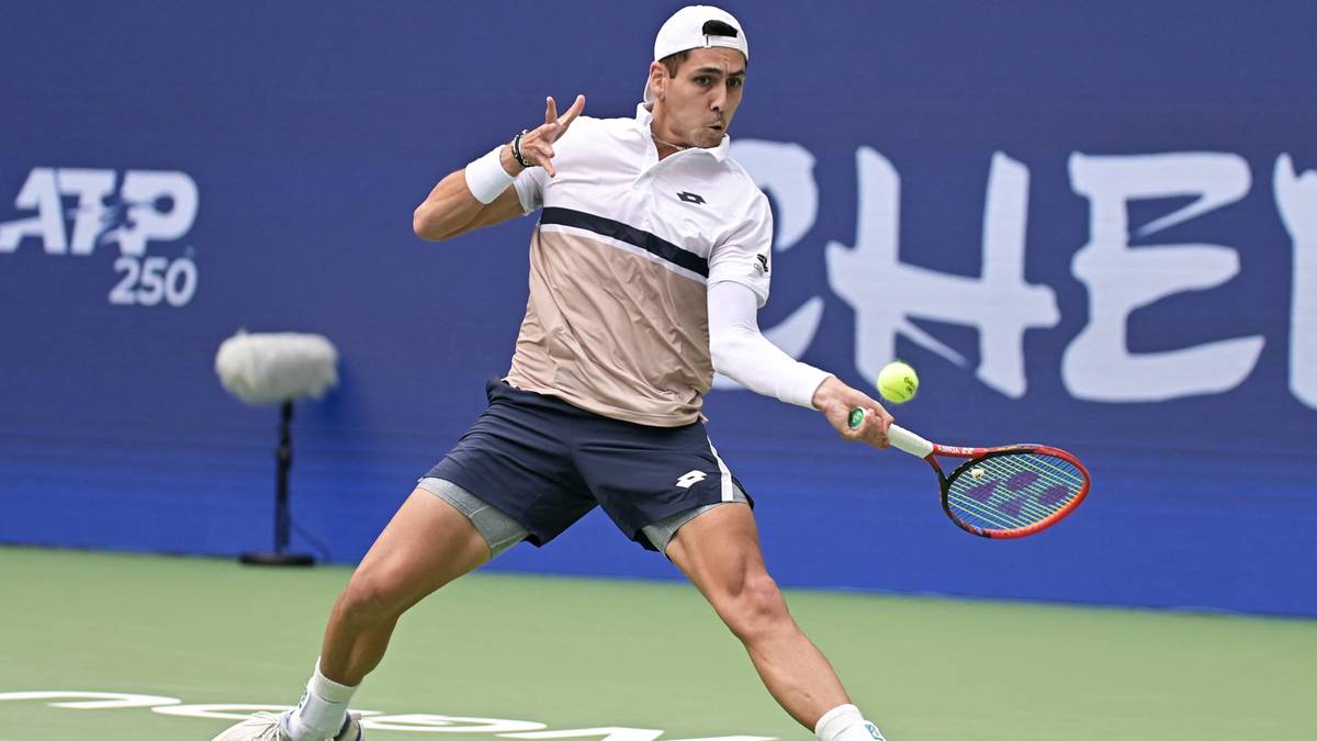 Tenisista w białej czapce z daszkiem, białej koszulce z ciemnymi pasami i granatowych spodenkach odbija piłkę rakietą podczas meczu.