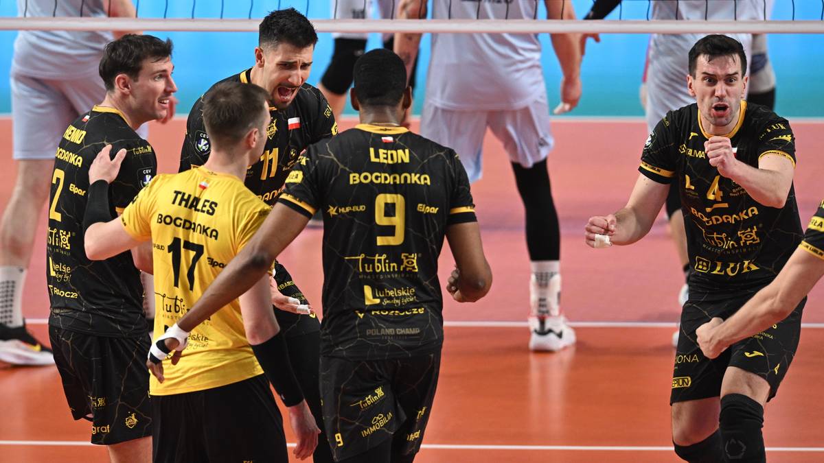 Grupa mężczyzn w strojach sportowych do siatk&oacute;wki, niekt&oacute;rzy w ż&oacute;łtych, inni w czarnych z ż&oacute;łtymi elementami, rozmawiają ze sobą na boisku.