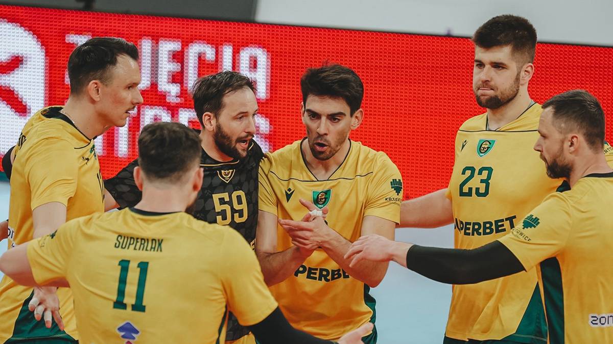 Sześciu siatkarzy w ż&oacute;łtych koszulkach z zielonymi akcentami i ciemnymi spodenkami, otacza się w kole na parkiecie hali sportowej. Mają na sobie ochraniacze na kolana i przepaski na głowach. Dw&oacute;ch zawodnik&oacute;w z numerami 11 i 59 jest odwr&oacute;conych plecami do widza, a pozostali patrzą na siebie.