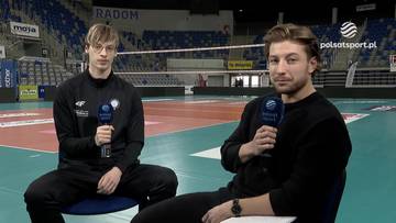 Dw&oacute;ch młodych mężczyzn siedzi na krzesłach na tle hali sportowej z boiskiem do siatk&oacute;wki. Obaj trzymają mikrofony.