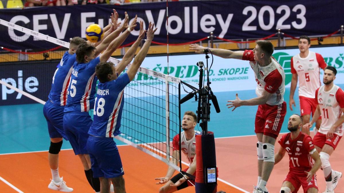 Siatkarze w biało-czerwonych strojach grają w piłkę siatkową przy siatce. Jeden z zawodników po prawej stronie wykonuje zagranie ręką nad siatką.