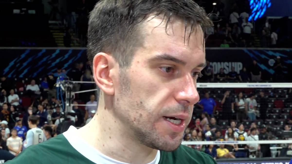 Siatkarz w bliżej nieokreślonym zielonym stroju sportowym, w zbliżeniu, z otwartymi ustami wypowiadający się. Na drugim planie widać trybuny wypełnione kibicami i oświetlenie hali sportowej.