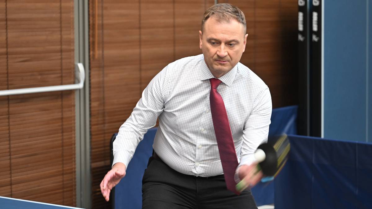 Mężczyzna w białej koszuli i bordowym krawacie gra w tenisa stołowego, uderzając piłeczkę rakietką.