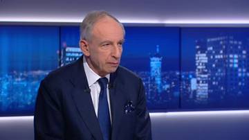 Mężczyzna w średnim wieku w granatowym garniturze i niebieskim krawacie siedzi w studiu telewizyjnym. Za nim znajdują się ekrany wyświetlające nocny widok miasta.