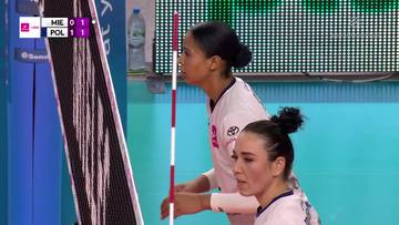 Dwie siatkarki w białych strojach meczowych, jedna z nich wykonuje zagrywkę, druga obserwuje grę. Na ekranie widać wynik meczu.