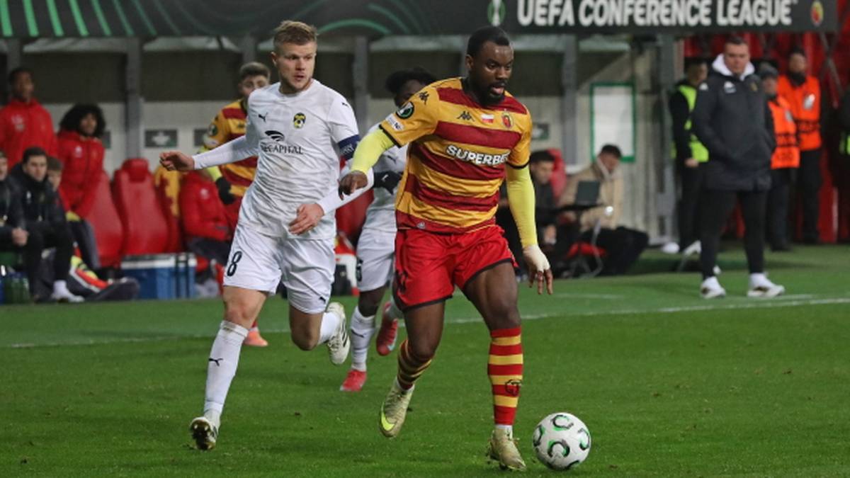 Dwóch piłkarzy podczas meczu piłki nożnej, jeden w żółto-czerwonym stroju prowadzi piłkę, drugi w białym próbuje go przejąć.