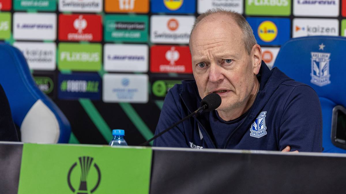 Mężczyzna siedzi przy stole konferencyjnym, przemawiając do mikrofonu. Za nim na ścianie widnieją kolorowe logotypy sponsorów.