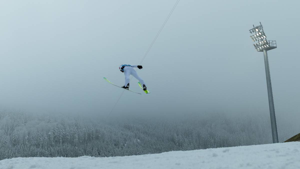 Skoczek narciarski w białym kombinezonie leci w powietrzu nad zaśnieżonym zboczem, w tle gęsta mgła i maszt oświetleniowy.