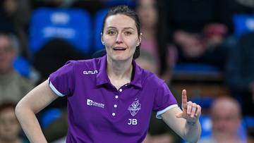 Kobieta w fioletowej koszulce z napisem JB, w okularach i z podwiniętymi rękawami, wskazuje palcem i uśmiecha się.