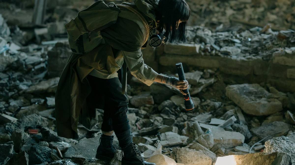 Osoba w masce gazowej i mundurze, trzymająca latarkę, przeszukuje zrujnowane, kamienne ruiny.