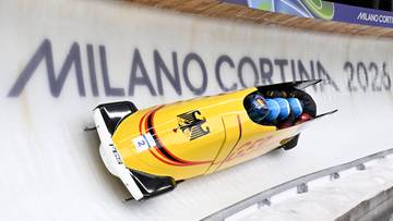 Ż&oacute;łty bobslej z niemieckimi oznaczeniami na lodowym torze, z napisem "MILANO CORTINA 2026" w tle.