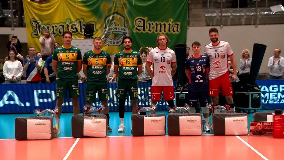 Sześciu siatkarzy w zielonych i białych strojach sportowych stoi na podium, trzymając nagrody. W tle widoczne są banery z logo i nazwami drużyn oraz publiczność.