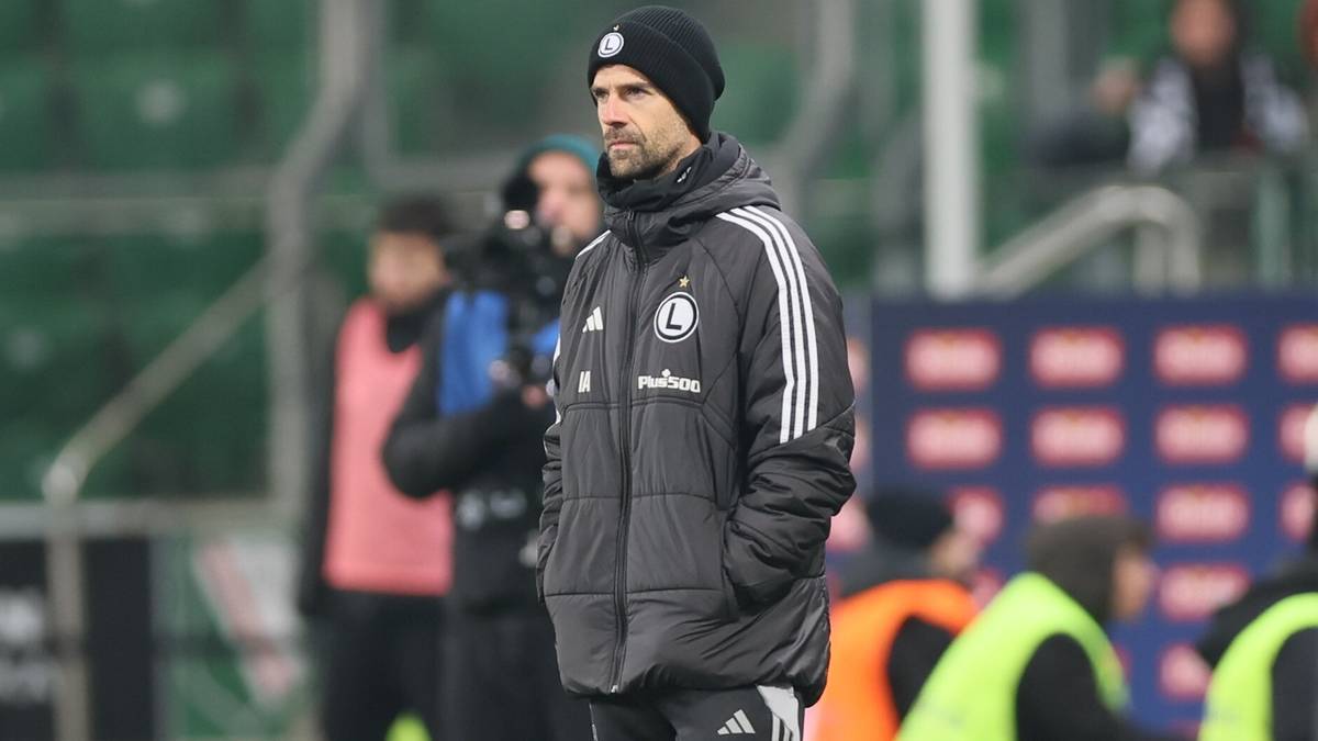 Trener Legii Warszawa w kurtce i czapce na boisku.