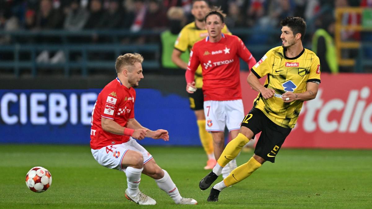 Dwóch piłkarzy na boisku, jeden w czerwonej koszulce próbuje odebrać piłkę zawodnikowi w żółtej koszulce.