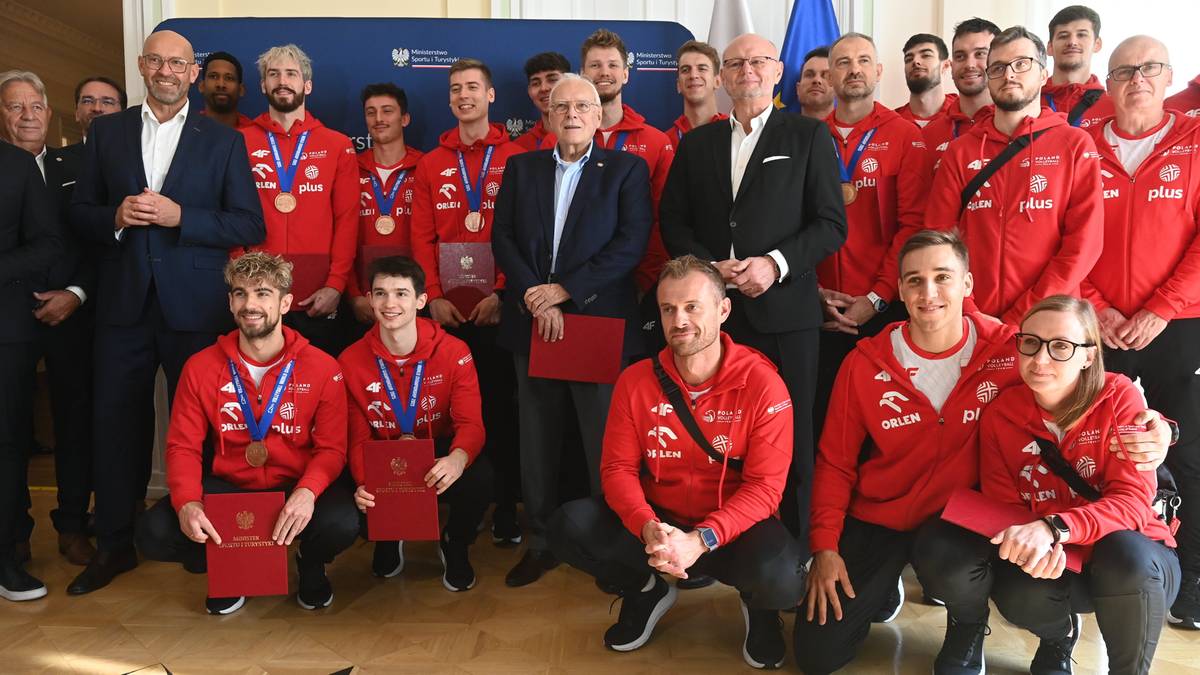 Grupa mężczyzn w czerwonych bluzach z kapturem, część z nich trzyma czerwone dyplomy lub pudełka. W środku stoi mężczyzna w garniturze z odznaką. Na ścianie w tle widnieje logo Ministerstwa.